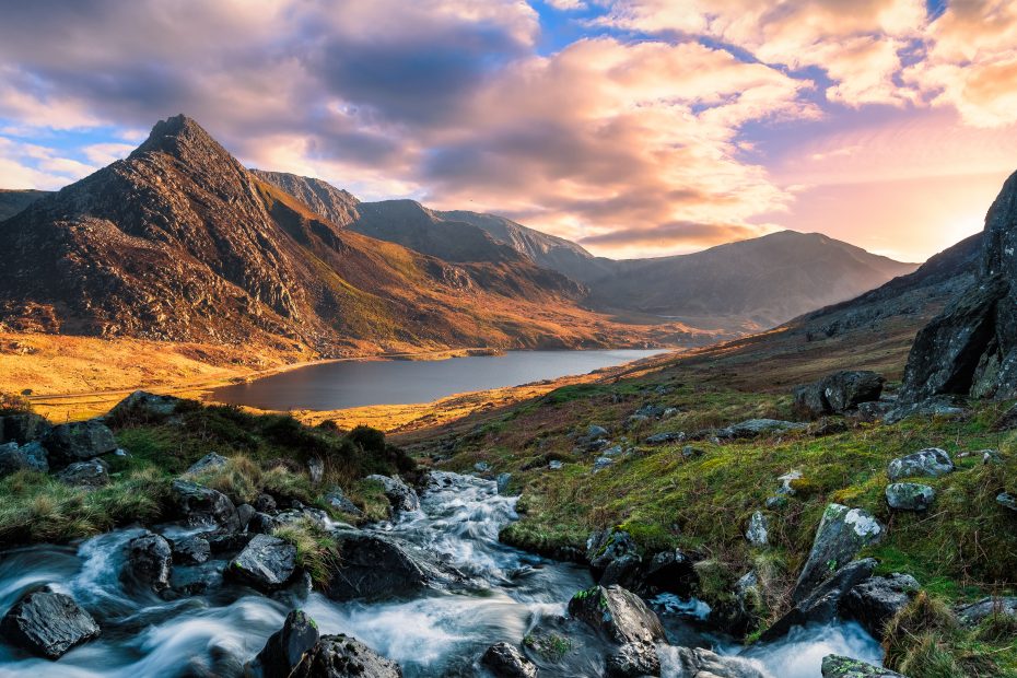 landscape in snowdonia national park with a stream in the foreground running towards a still lake at the foot of a mountain near sunset with pink clouds overhead in wales