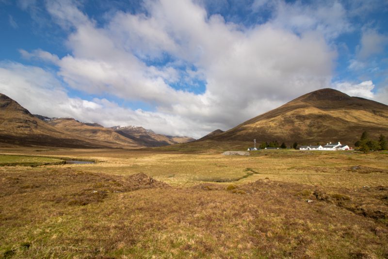 Heather and moorland with mountains in the distance and the white painted building of Cluanie Inn beneath the right hand mountain