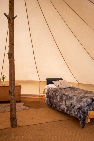 interior of a white canvas bell tent with a single bed with grey fluffy throw on top of it and a large wooden pole in the centre holding the tent up, there is a beige fluffy rug on the floor and a small wooden table with a drawer