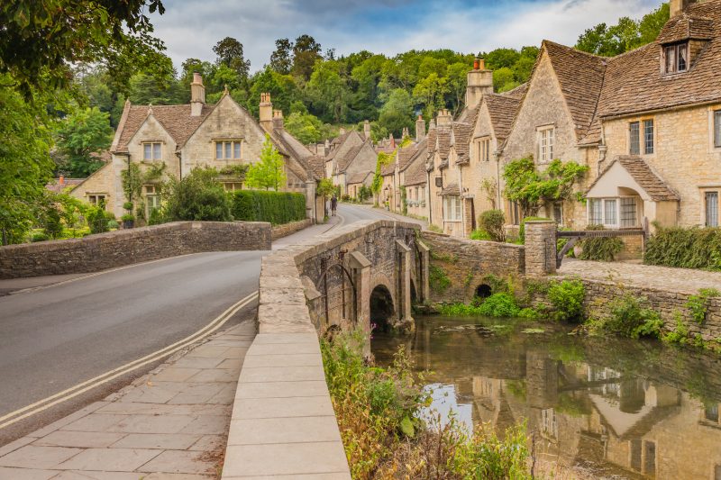 a stone bridge with a narrow road over a shallow calm river and a row of beige stone cottages behind and a green woodland behind that - tips for driving in the cotswolds