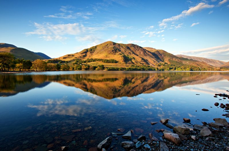 a very still lake surface reflecting a tall green hill and blue sky at Buttermere in the Lake District England 