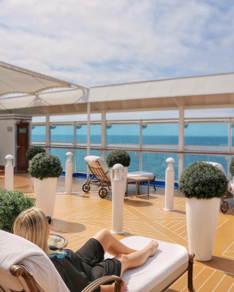 Emily wearing a long black kaftan relaxing on a white sunbed in the Sanctuary on the Regal Princess, the bed is outside on the deck of the ship with empty wooden floor in front dotted with large white plant pots with green leafy hedges. Emily is facing a glass wall with the sea beyond. 