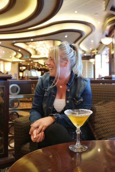 Emily wearing a white top and a blue denim jacket with her blonde hair in a messy ponytail looking to the side and laughing whilst sitting on an armchair in a bar with a cocktail in front of her in a triangular glass with sugar around the rim. 