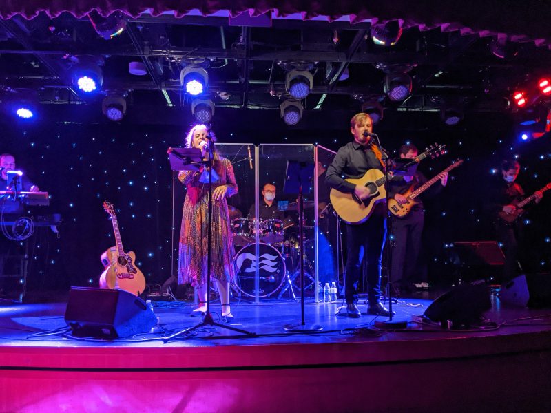 A man holding a guitar and a woman in a dress singing on stage with a band behind them - entertainment on the Regal Princess