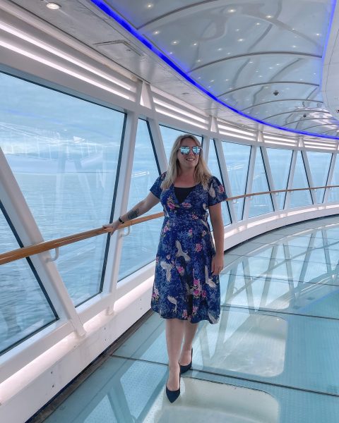 Emily wearing a mid-length blue silk dress with white and pink birds on it, black high heels, and purple sunglasses with her blonde hair down, walking along a glass walkway in a glass tunnel on a cruise dhip with the sea underneath her and out the window behind her.