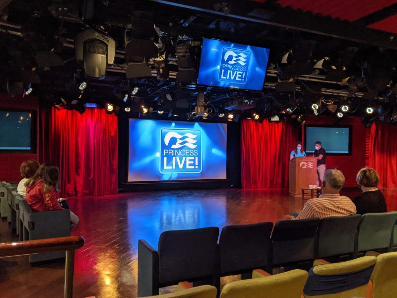 theatre stage with two large TV screens with a blue background and the words Princess LIVE! written on them in white, the stage has red curtains on either side and there are two people wearing face masks standing at a wooden lecturn. 