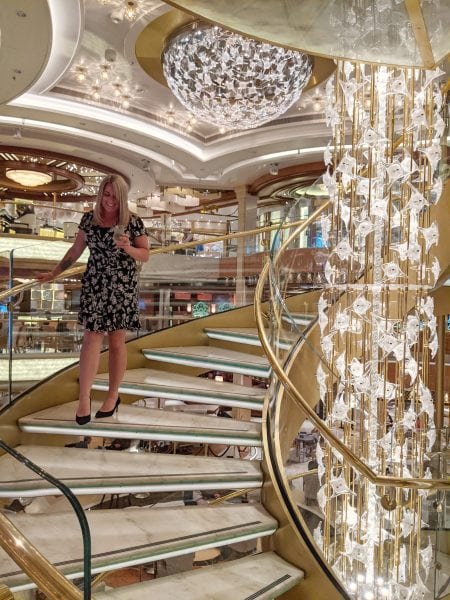 Emily walking town a marble spiral staircase with gold banisters and a gold central column covered in fairylights in the Atrium of the Regal Princess cruise ship