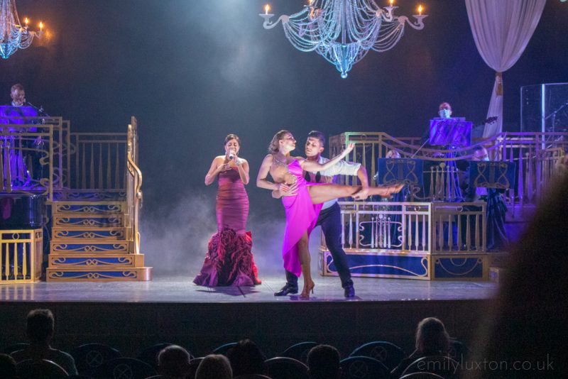 stage in a theatre with a woman wearing a long red sleavless ball gown with a ruffled skirt holding a microphone and singing next to a couple dancing with the woman wearing a magenta dress and the man wearing a white shirt and black trousers, the woman has one leg up with the man holding it. the stage is black with dry ice lit up by spotlights and two chandeliers above. Entertainment on the Regal Princess