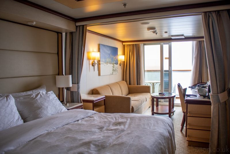 Interior of a cruise ship suite cabin with cream walls. there is a double bed near the camera with a white bedspread and a bedside table with a white lamp next to it. beyond that is a small lounge area with a beige three seater sofa, a wooden coffee table with a glass top, and a wooden desk with a wooden chain with a white back. beyond that is a set of French doors slightly open with the ocean just visible through the gap. Regal Princess Mini Suite cabin review