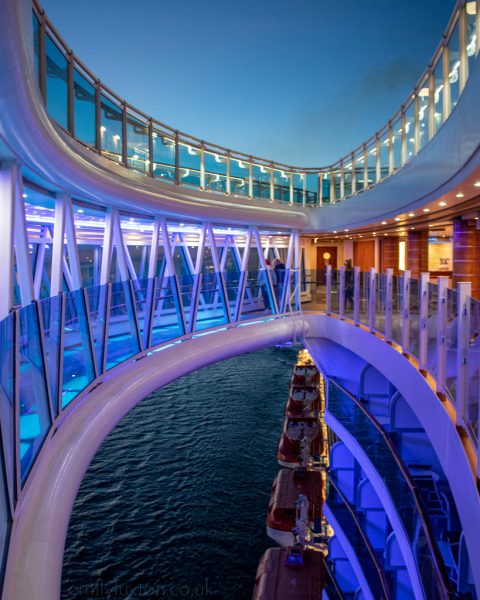 Regal Princess Seawalk at Night, a curved covered walkway made of glass with a white frame and glass floor. the walkway curves away from the side of the ship creating an oval space in between and the camera is looking through this with the ocdean and part of the ship deck down below. the walkway is lit up in purple against the night sky. 