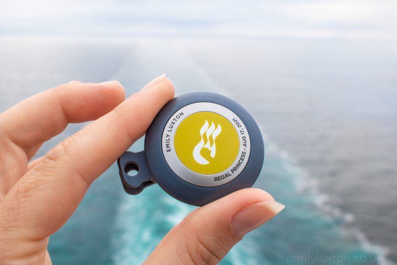 Close up of a hand holding a small grey circular badge with a yellow centre and the name Emily Luxton printed on it held in front of the ocean. Princess Cruises OceanMedallion