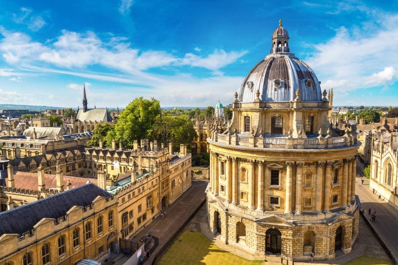 Radcliffe camera building in Oxford, a cicular three storey building bult from beige stone with a grey domed roof, with a row of terraced beige stone buildings alongside and the city skyline of oxford visible behind on a sunny day with blue sky above