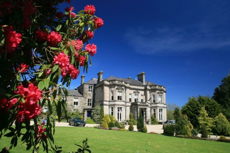 exterior of a large grey stone country manor hotel with a grassy lawn in summer framed by a bush of pink flowers