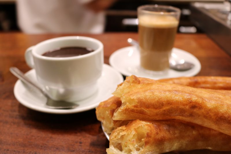 large churros on a wooden bar next to a white mug of chocolate sauce at El Comercio in Seville