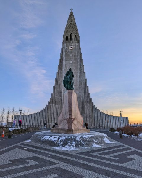 a church with a tall pointed tower behind a stature of a man with a small patch of snow on the ground and sunset behind the church in Reykjavik