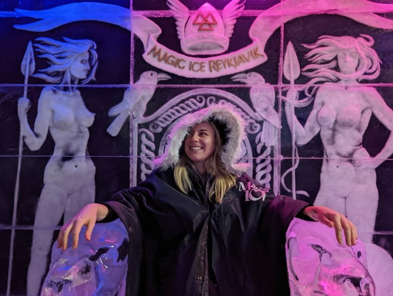 Emily wearing a black cape with fur hood sitting in a throne made of ice under a sign saying Magic Ice Bar in Reykjavik