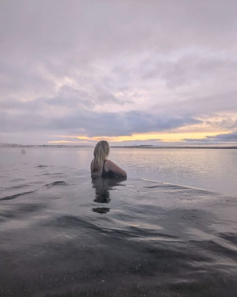 Emily reclines on the edge of the infinity pool at Iceland's Sky Lagoon, overlooking the sea under a gray sky with a faint glow at dawn