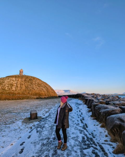 Emily in a pink hat and long grey winter coat standing on snow in front of a hill next to Reykjavik Harbour