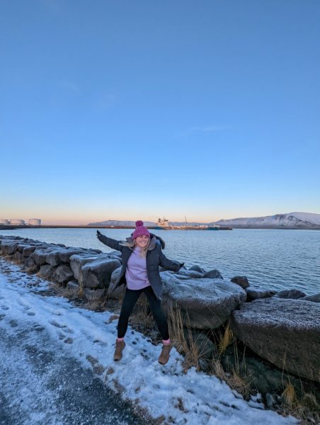 Emily wears a long purple jumper and pink winter hat and is jumping ona snowy ground in front of some rocks with the sea in the background in reykjavik