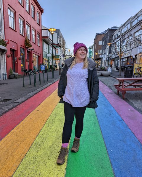 Emily wearing a purple jumper, pink hat and grey winter coat standing on a street painted in Rainbow stripes for Pride in Reykjavik