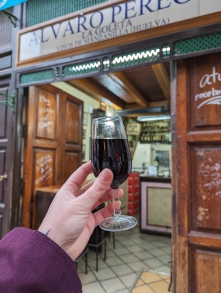 hand holding a small glass of dark red wine in front of the open door of a small bar in Seville