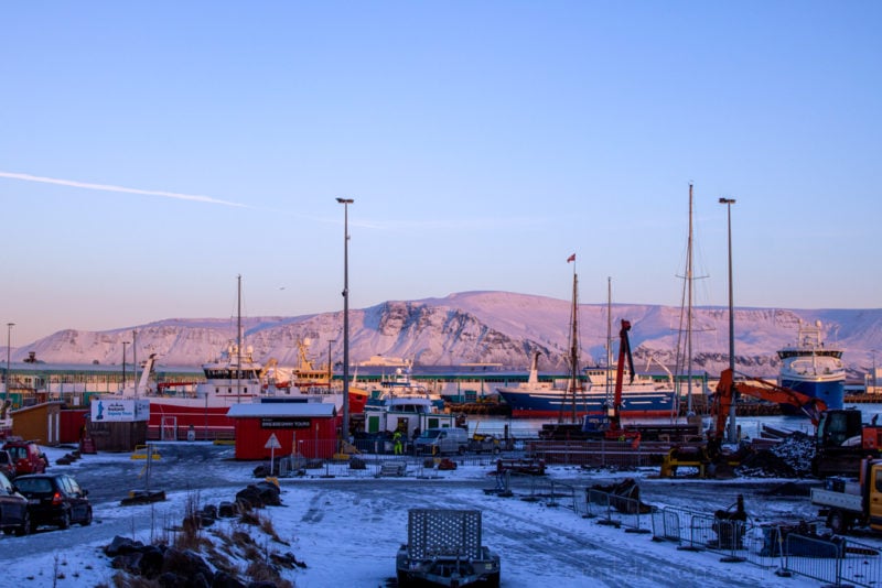 lots of boats and tall hip masts in front of a large snow covered hill with snowy ground at Reykjavik Harbour in winter
