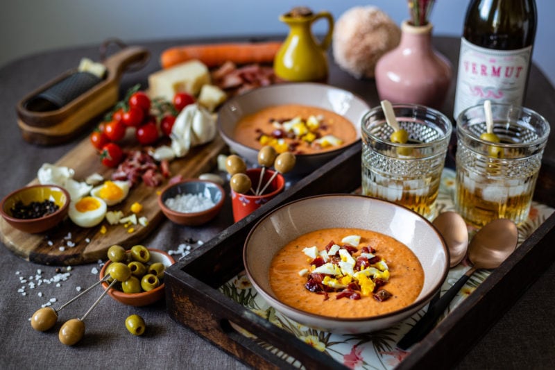 a table crowded with different traditional seville food dishes including bowls of soup, ham, olives, and glasses of vermouth