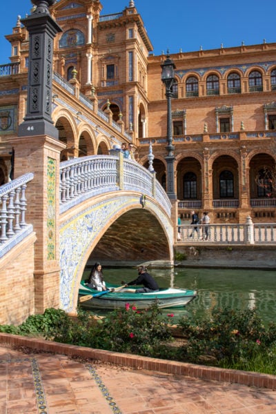 A turquoise rowing boat emerging from under a bridge with an orange briick palace in the background in the Plaza de Espana Seville
