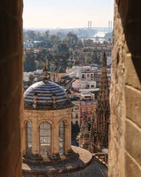 Looking out a narrow window in a stone tower with a view of a domed tower and a city in the background