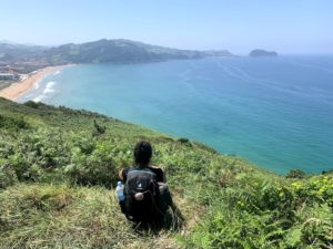 woman with a backpack on sitting on the edge of a grassy cliff with a view of the ocean
