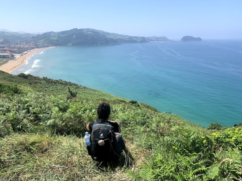 girl with dark hair wearing a backpack sitting on a cliff edge overlooking the sea