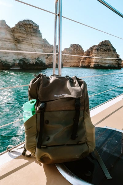 Close up of a dark green travel backpack on the edge of a boat with the sea and rocks behind it