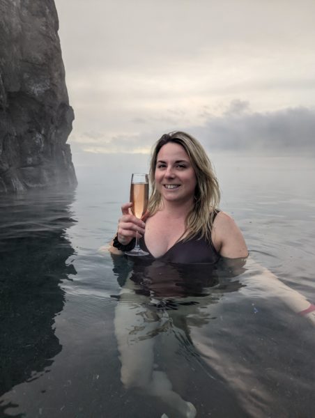 Emily wears a purple swimsuit, holds a glass of sparkling wine and smiles at the camera in a pool with gray cliffs behind.