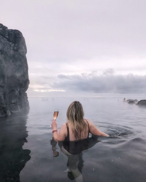 back of a girl in a swimsuit with long blonde hair holding a glass of pink sparkling wine in a pool with grey sky behind