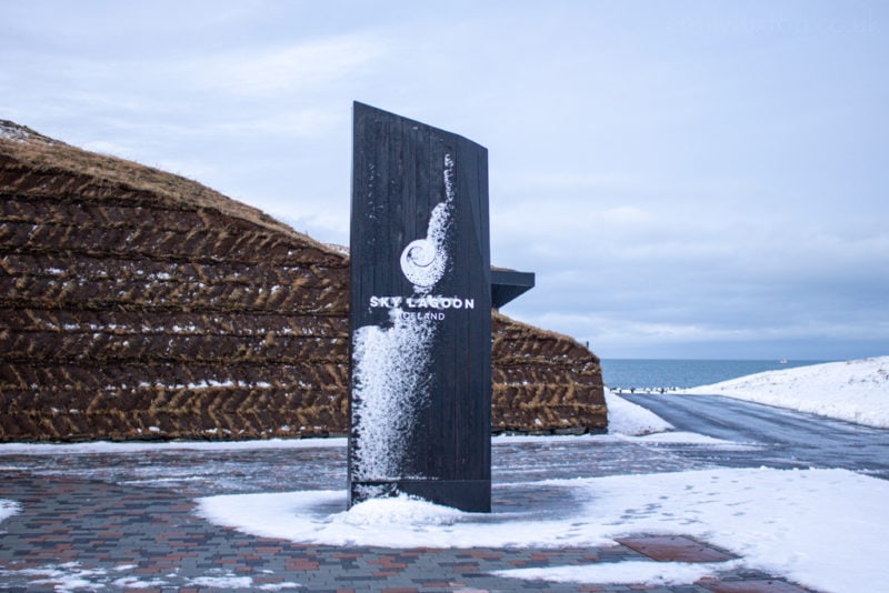 snow covered sign which says Sky Lagoon Iceland in front of a low wall made from earth and grass with snow and the sea in the background