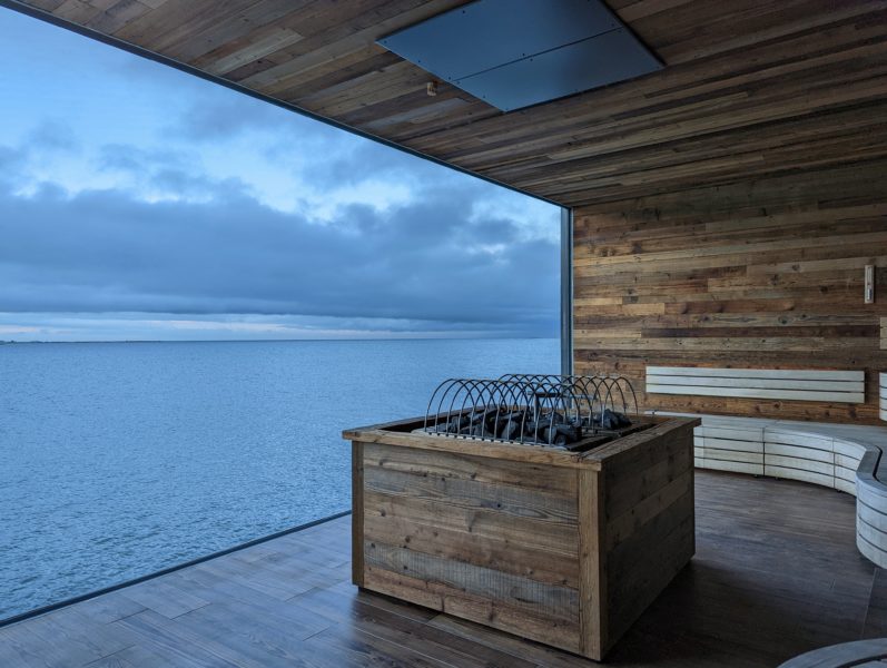 wooden interior of a sauna with a square coal heater in front of a huge glass window overlooking the ocean on a grey day