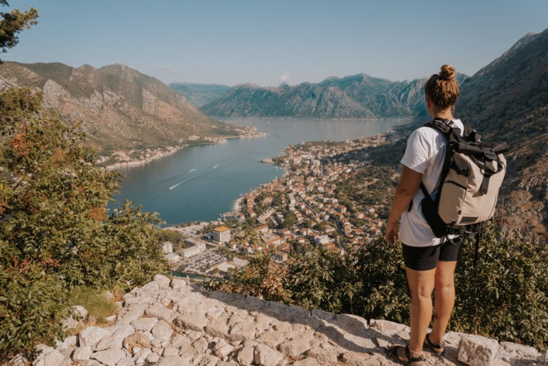 a girl wearing a white t shirt with a small grey backpack standing on a hill looking over a lake and a small town - best carry on bags for women
