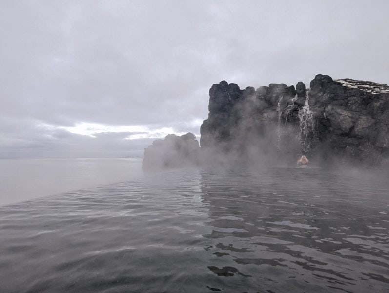 grey water with steam rising on it in front of a black rocky cliff with a small waterfall