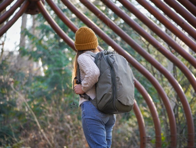 girl wearing a white jumper, jeans and an orange hat with a large grey carry on backpack standing in front of trees looking away from camera