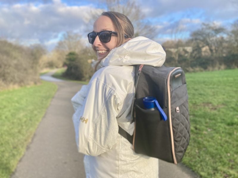 girl wearing a white raincoat and black carry on backpack turning to look at the camera on a path in a park