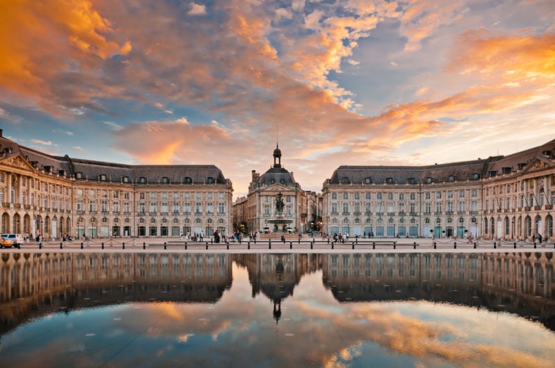 a  grand old building with a grey roof in the centre of Bordeaux reflected in a pool of water at sunset