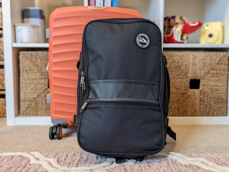 black carry on backpack in front of an orange suitcase and a bookshelf, it is the Cabin Max Chicago, one of the best carry on bags for women