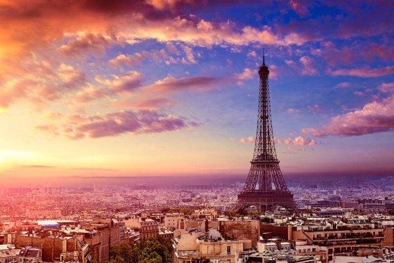 view of Paris city skyline with the Eiffel Tower rising above the buildings against a pink and purple sky at sunset. 