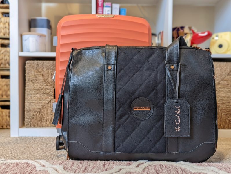 black tote bag in front of an orange suitcase with a bookshelf in the backgroun, the bag is the Travel Hack tote bag, one of the best carry on bags for women