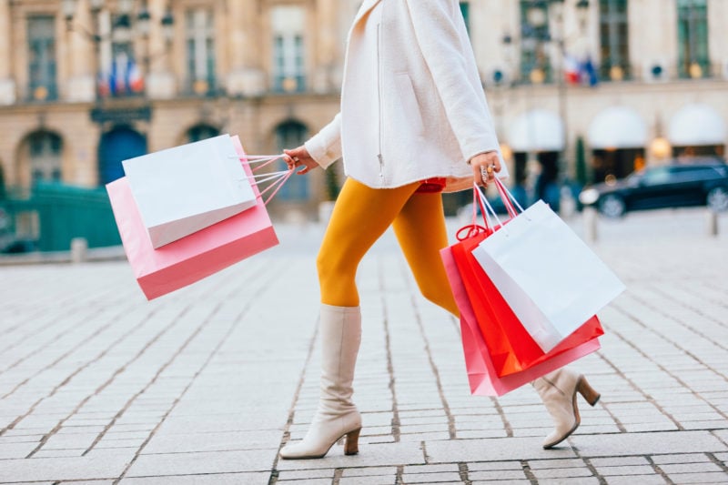 bottom half of a woman wearing a white jacket, yellow tights and white knee high boots, walking through Paris while holding many paper shopping bags 