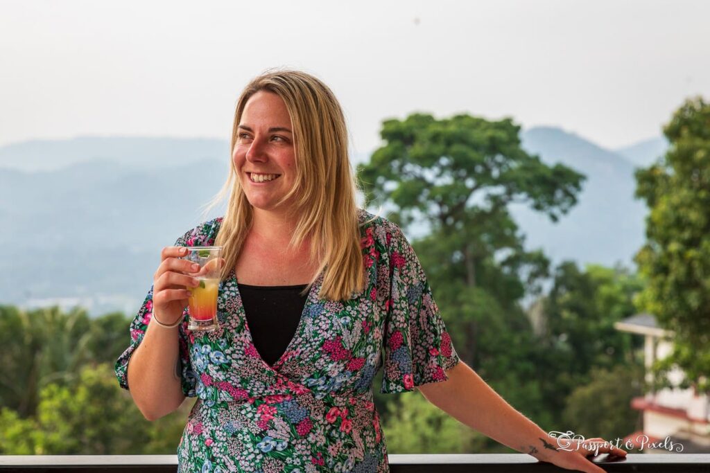 Close up of UK solo female travel blogger Emily wearing a green and pink dress with a floral pattern and holding a glass of an orange coloured cocktail. she is smiling and looking away from the camera and there is a view of hills and jungle trees out of focus behind her.