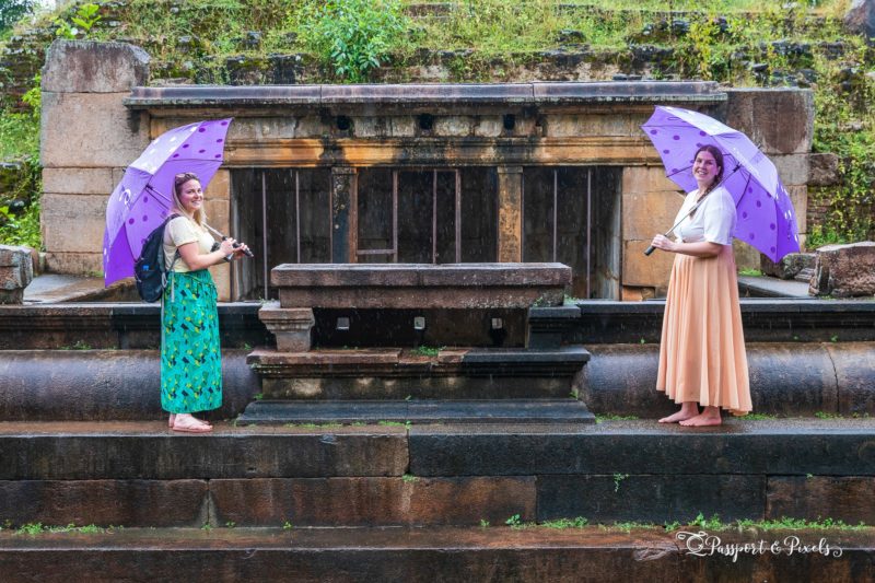 emily wearing a long green skirt and yellow t shirt facing lisa wearing a peach coloured skirt and white top. both are holding purple umbrellas and standing on a stone wall with an ancient ruin behind. 