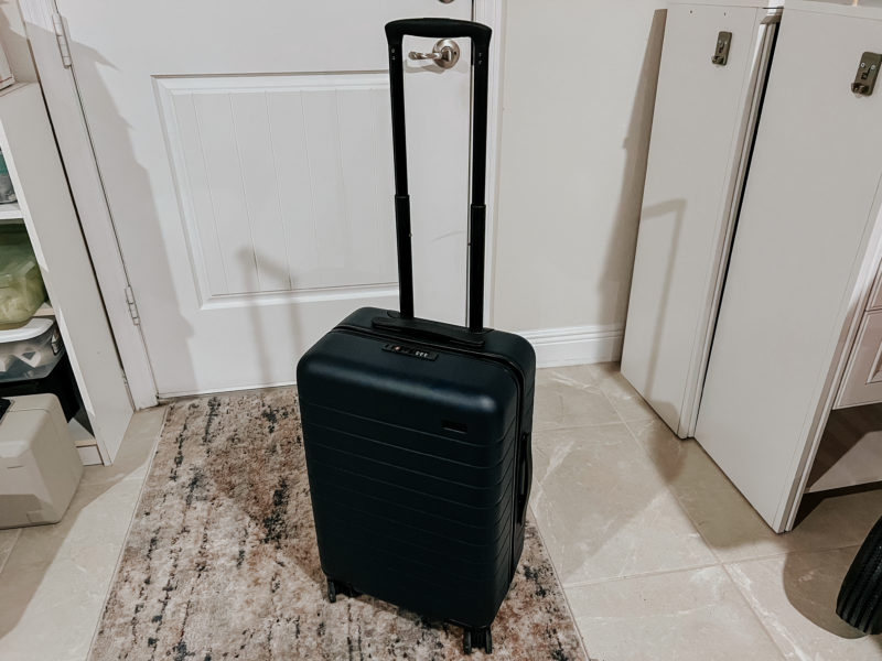 Small black suitcase with handle extended in front of a white door - Away Carry On luggage for women