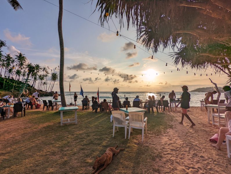 silhouettes of many people sitting at bar by the sea in front of a sunset