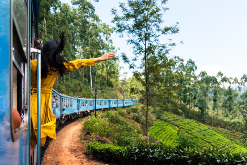 woman with black hair and a yellow dress leaning out of the door of a blue train as it passes a tea plantation - adventures in sri lanka